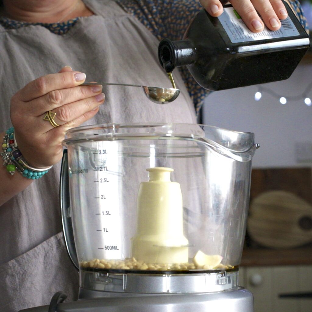 Woman pouring oil from a brown bottle into a silver measuring spoon over a food processor bowl