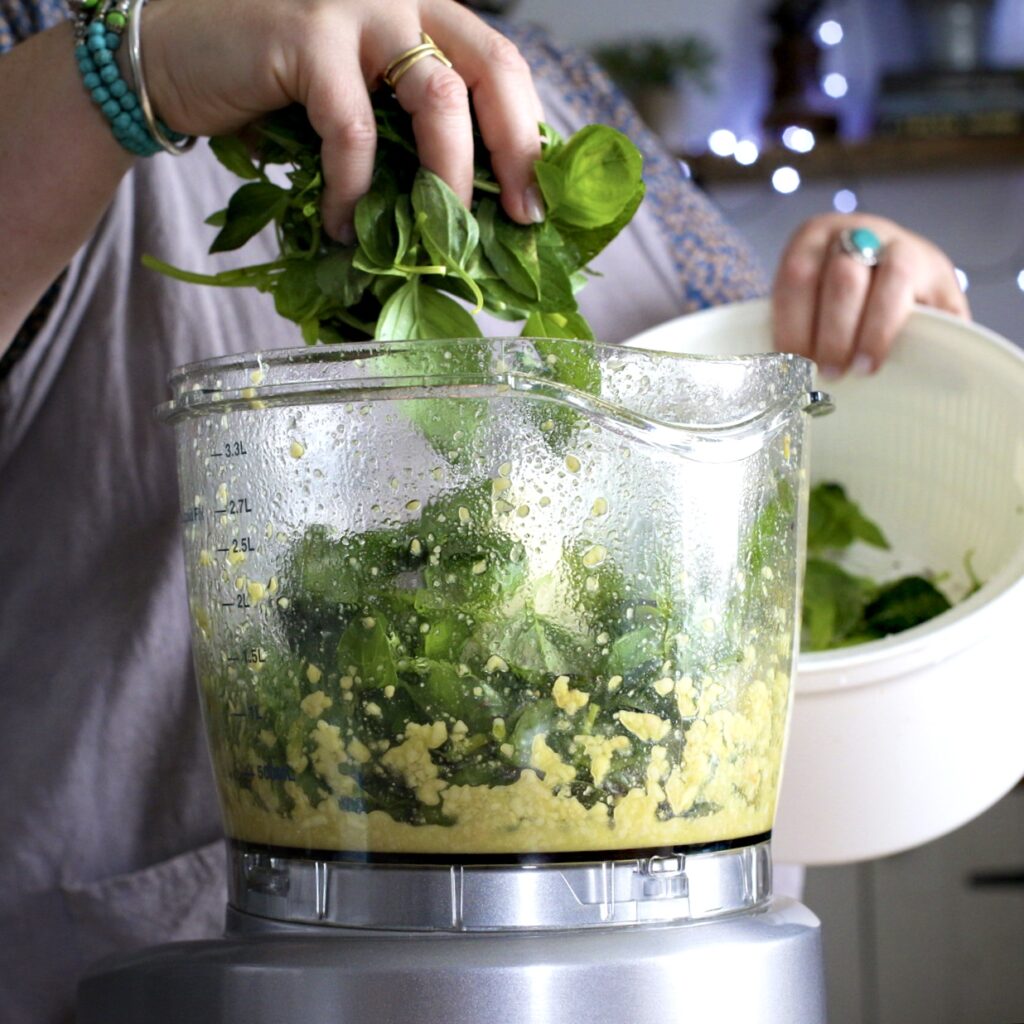 Woman in grey placing handful of bright green basil into a food processor bowl