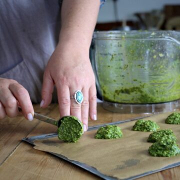 Womans hands placing small piles of fresh homemade pesto onto a baking tray with a silver tablespoon next to a mixing bowl of homemade pest