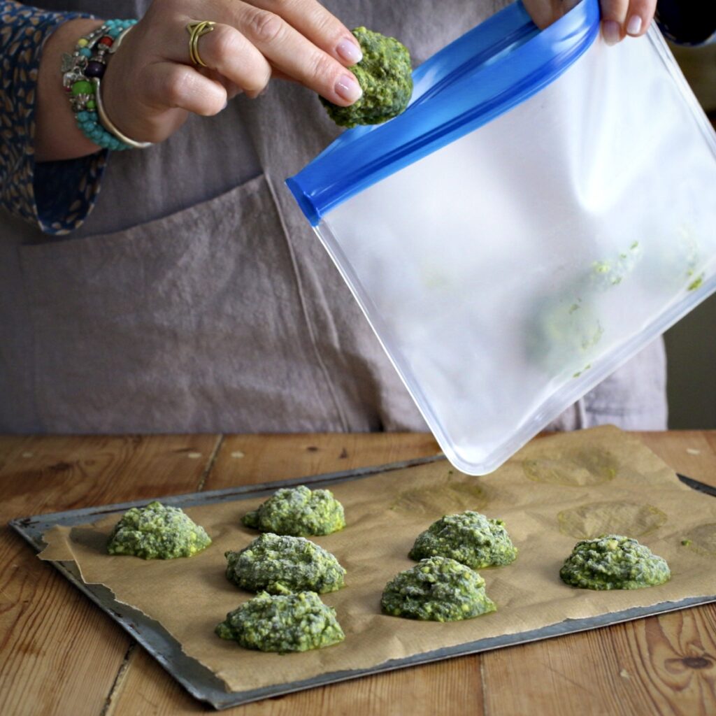 Womans hands placing a frozen a small scoop of frozen pesto into a silicone freezer bag