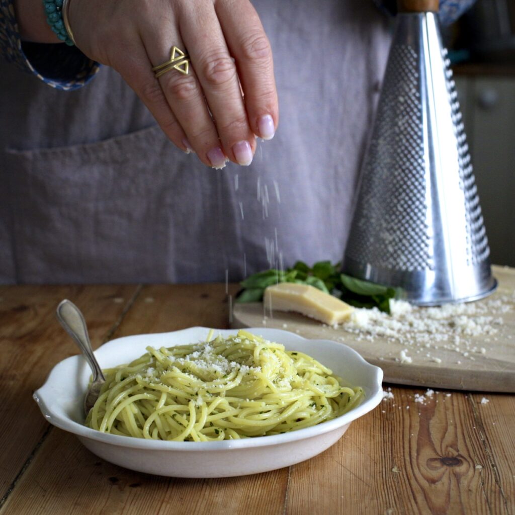 Womans hands sprinkling grated Parmesan over a white bowl of pesto spaghetti