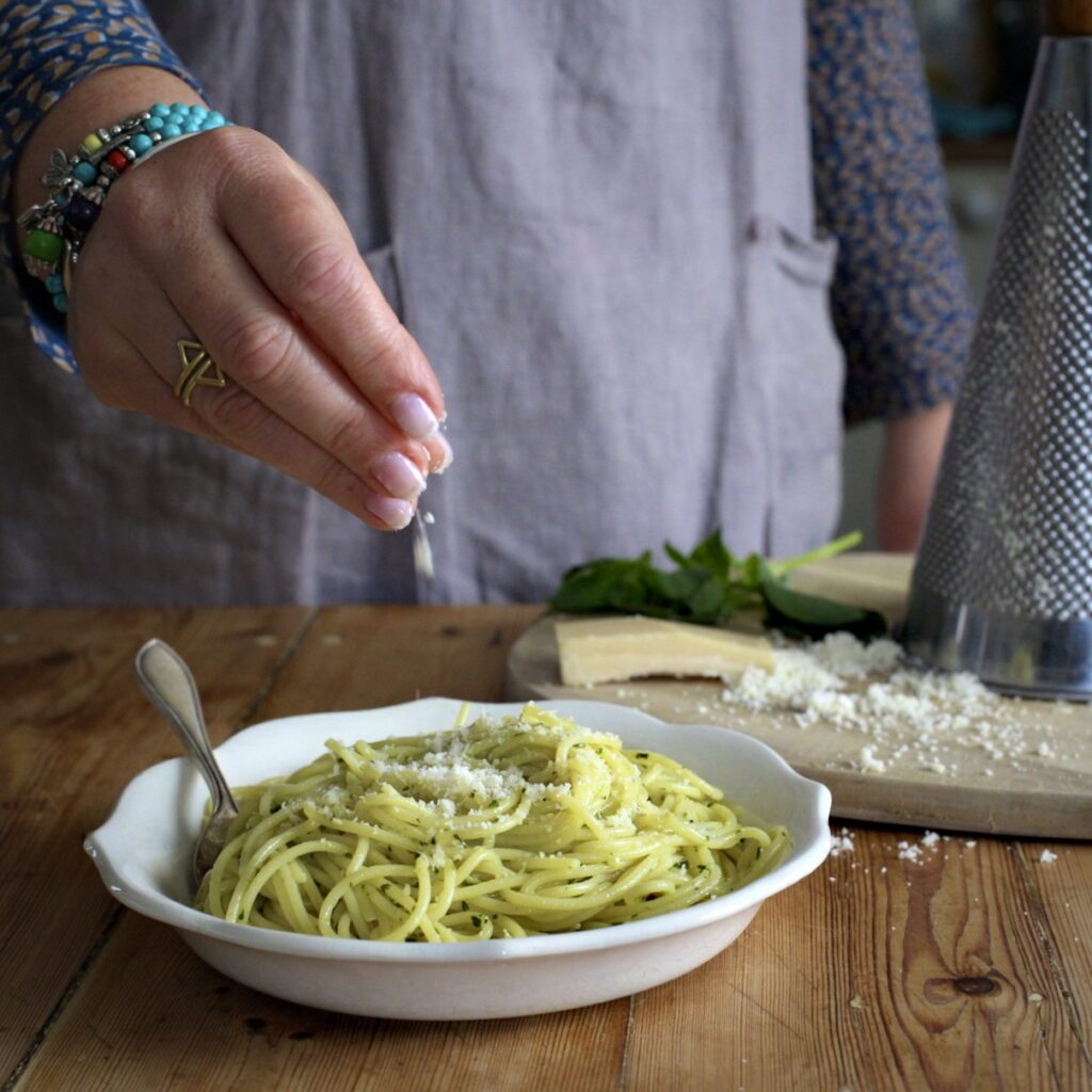 Womans hands sprinkling grated Parmesan over a white bowl of pesto spaghetti