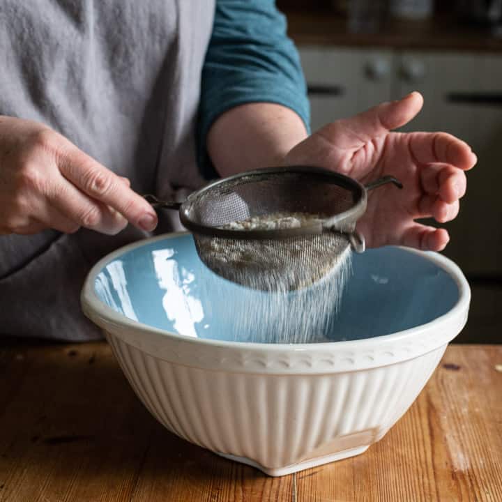 womans hands sieving flour into a blue mixing bowl on a wooden kitchen counter