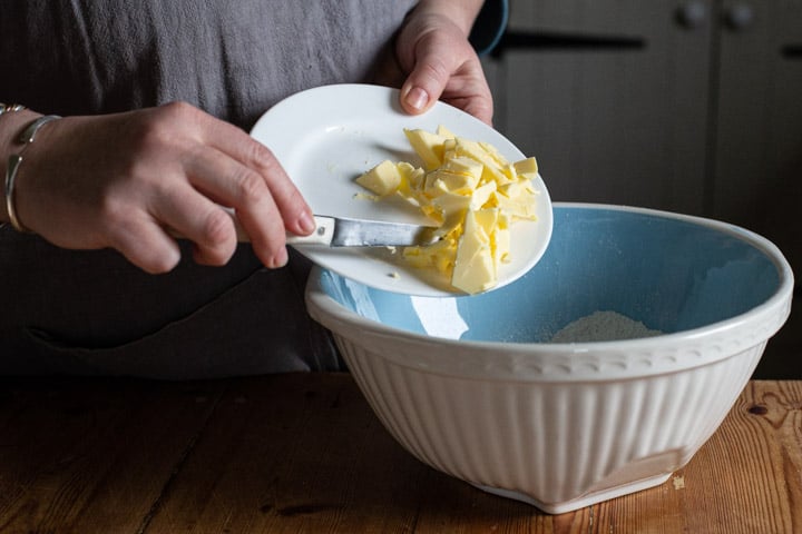 womans hands scraping butter from a white plate into a blue mixing bowl on a wooden kitchen counter