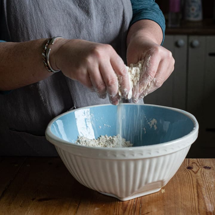 womans hands rubbing butter into flour in a blue mixing bowl on a wooden kitchen counter