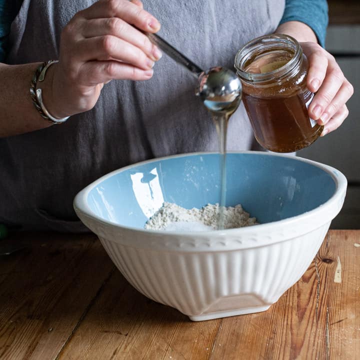 womans hands pouring honey from a silver spoon into a blue mixing bowl