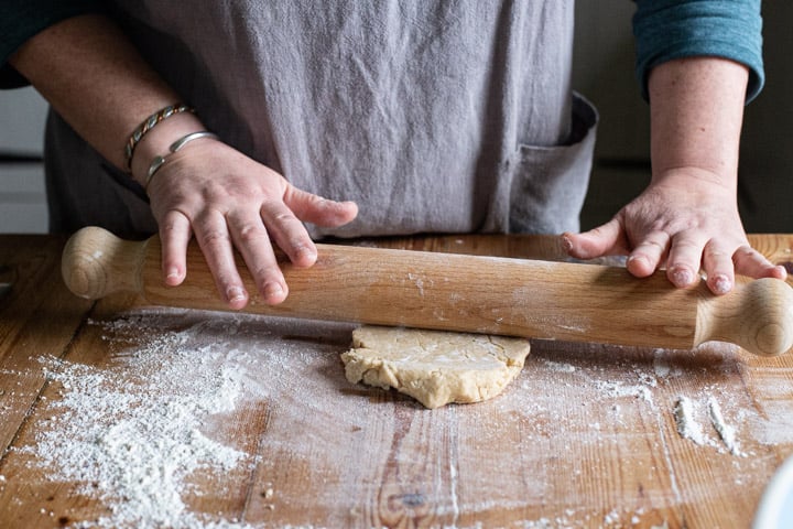 womans hands rolling cookie dough with a wooden rolling pin on a wooden kitchen counter dusted with flour