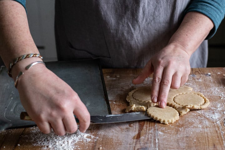 womans hands lifting a circle of cookie dough off a wooden kitchen counter with a silver palette knife 