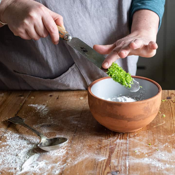 womans hands scraping bright green lime zest into a small bowl of icing sugar on a wooden counter