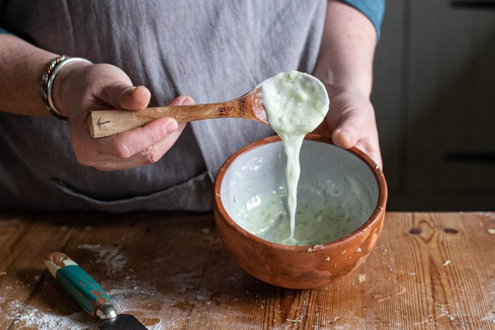 womans hands pouring white icing speckled with lime zest from a wooden spoon into a small terracotta bowl 