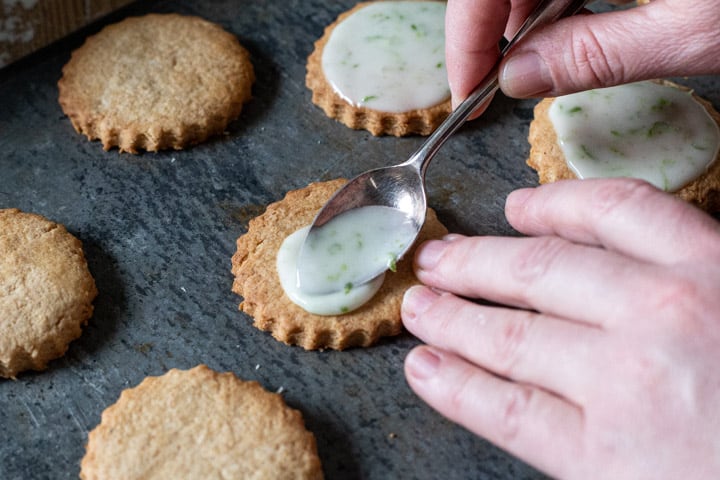 grey baking sheet with 5 cookies and womans hands spooning on white icing that is flecked with bright green lime zest