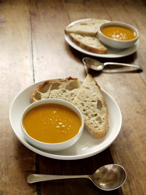 2 white bowls of Curried Sweeet Potato and Red Pepper Soup on white plates with bread and a soup spoons on a wooden table