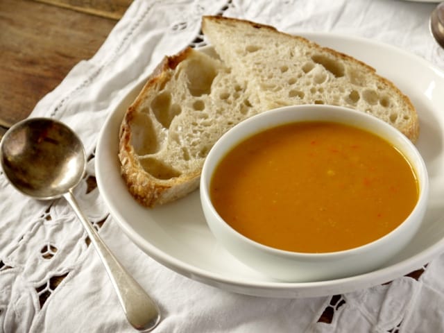 Curried Sweeet Potato and Red Pepper Soup in a whitebowl on a white plate with bread and a soup spoon on a lace cloth