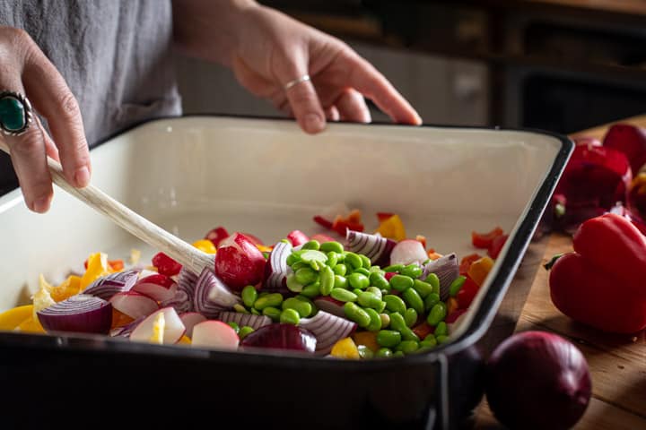 womans hands mixing together freshly roasted vegetables with hot couscous in a black and white roasting tin