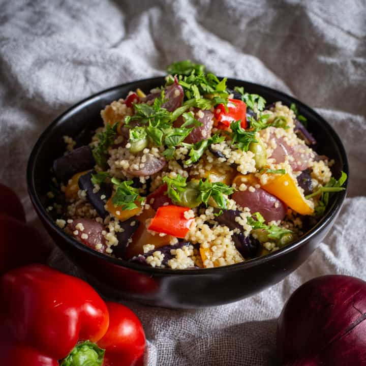 black bowl filled with colourful roast veg couscous against a natural fabric background with fresh veggies