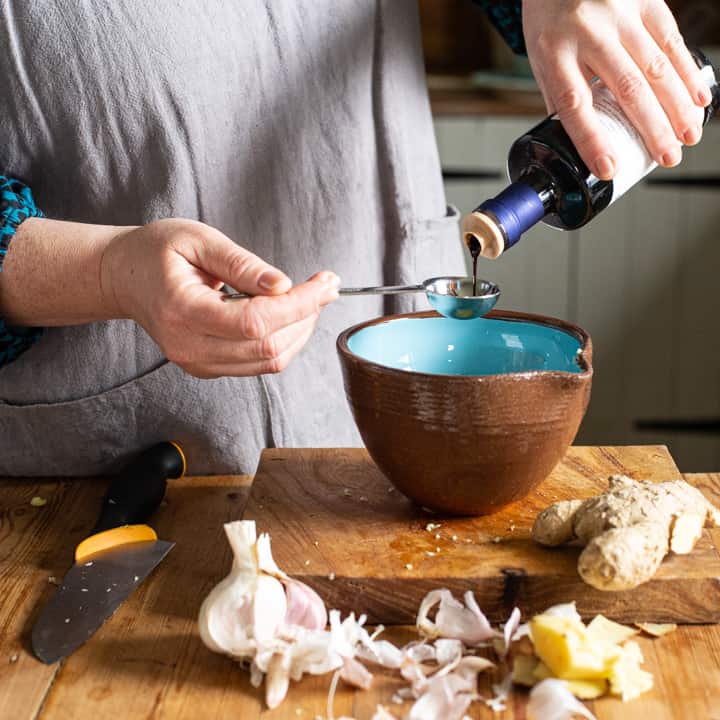 womans hands pouring balsamic vinegar from a black bottle into a silver measuring spoon over a blue bowl