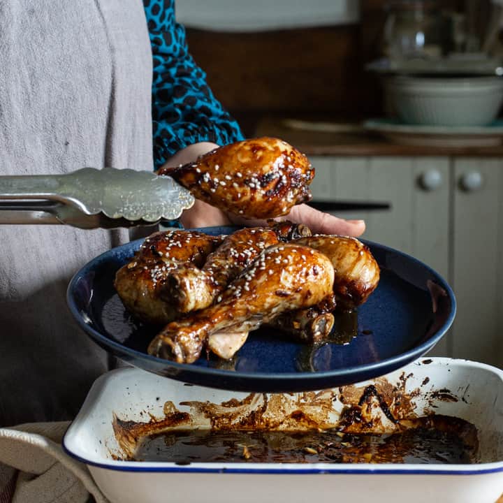 woman moving baked chicken drumsticks from a white baking tin onto a dark blue plate with a pair of silver tongs