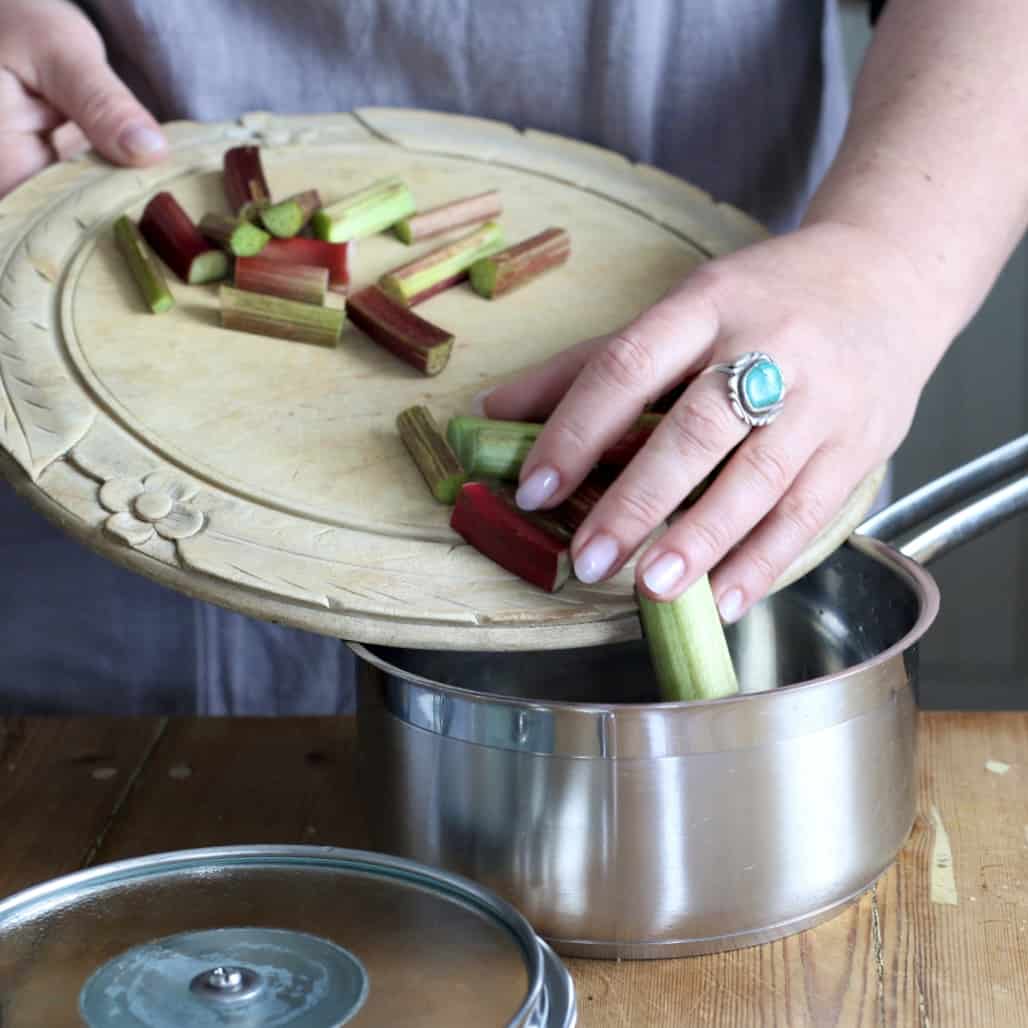Woman sliding cut pieces of raw rhubarb from a wooden chopping board into a silver saucepan