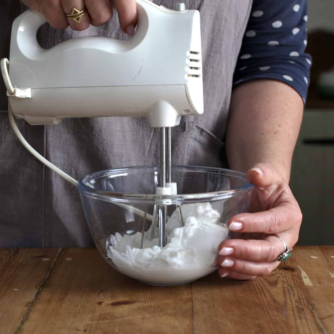 Woman in grey using a white electric whisk to beat canned coconut milk in a glass mixing bowl