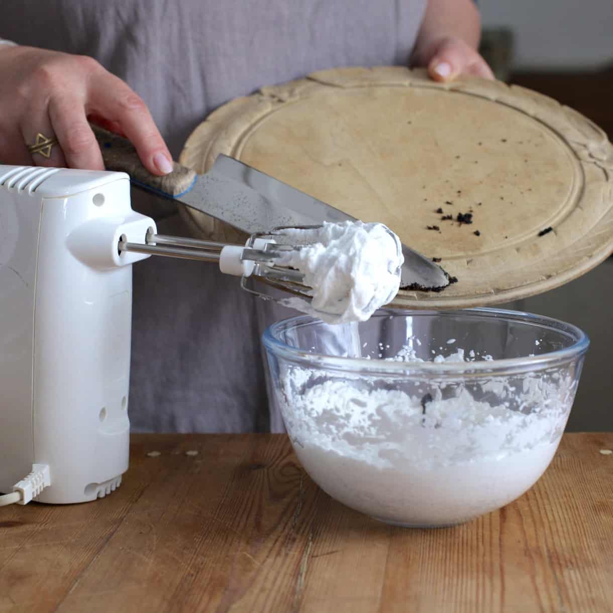 Hand mixer and glass bowl of whipped coconut cream with a woman scraping vanilla beans from a wooden board into the bowl of whipped cream