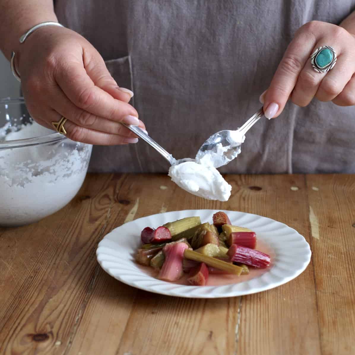 Woman spooning whipped coconut cream on top of a pile of poached rhubarb on a small white plate