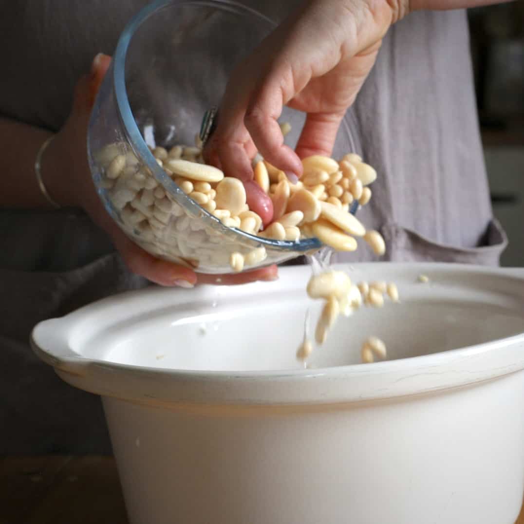 Woman tipping soaked butter beans into a white casserole dish form a small glass bowl