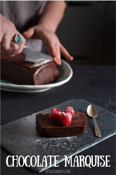 Slice of dark chocolate marquise cake on piece of slate with 3 raspberries and icing sugar on top