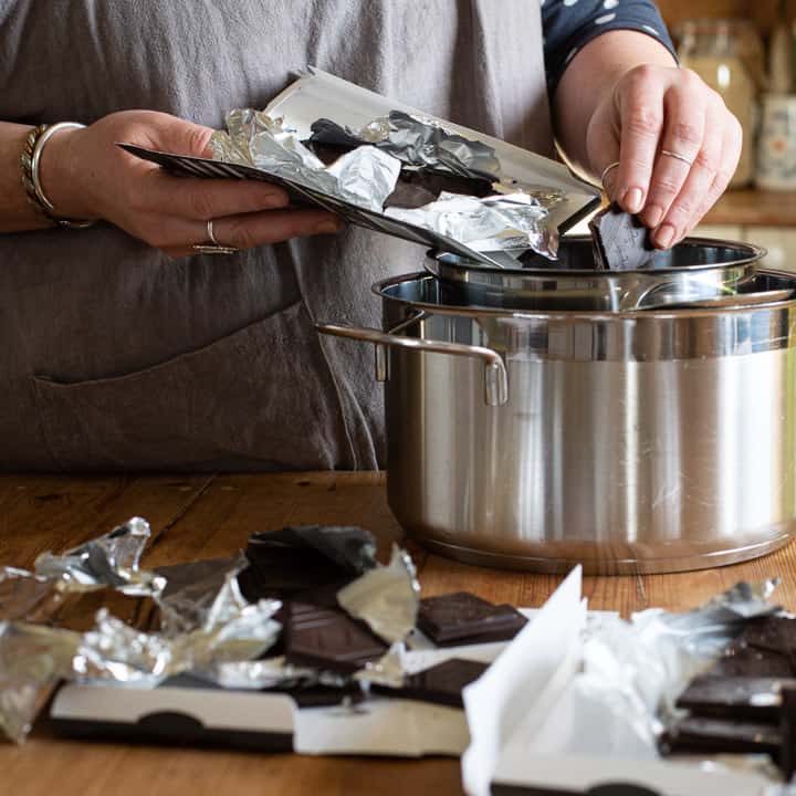 womans hands dropping broken pieces of dark chocolate into a silver saucepan 