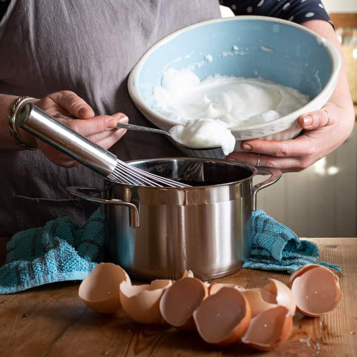 womans hands spooning frothy egg whites into a saucepan on a messy wooden kitchen counter