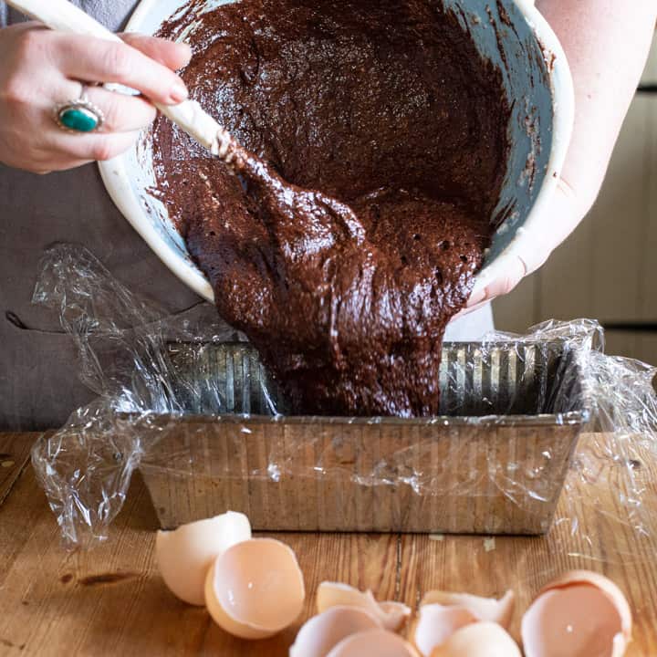 womans hands pouring chocolate marquise from a mixing bowl into a silver loaf tin on a messy wooden kitchen counter