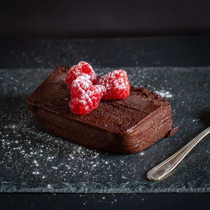 Slice of dark chocolate marquise cake on piece of slate with 3 raspberries and icing sugar on top