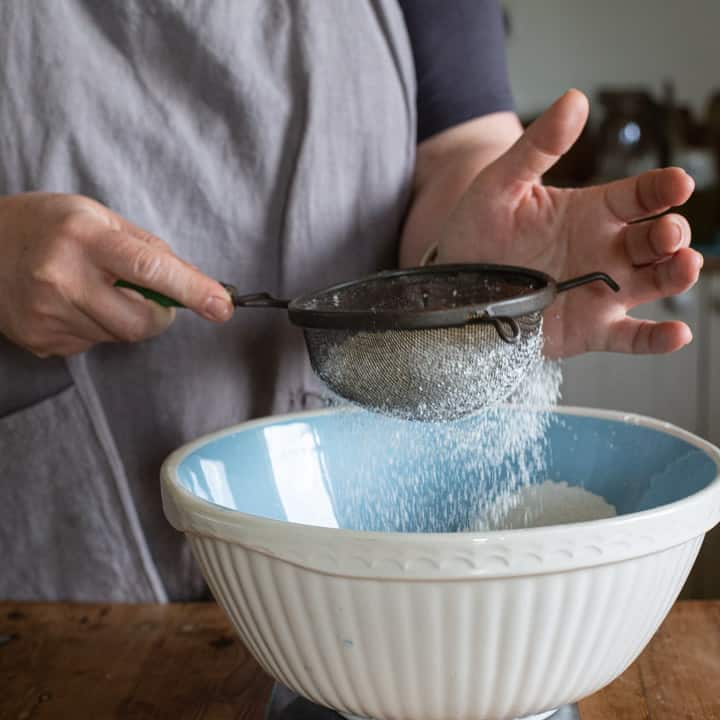 woman in grey sifting flour into a blue and white mixing bowl