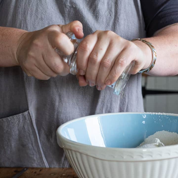 woman in grey grinding black pepper into a blue bowl of strawberry muffin batter