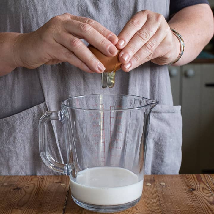 woman in grey cracking an egg into a glass jug of milk