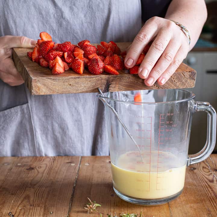 woman in grey sliding chopped fresh strawberries from a wooden chopping board into a glass jug of muffin batter