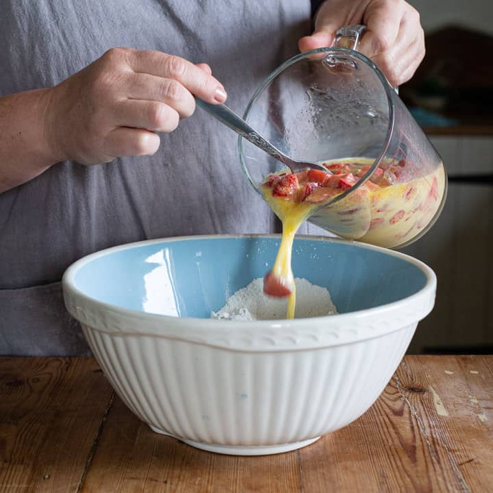 woman in grey pouring the a jug of wet muffin ingredients into a blue bowl of dry muffin ingredients