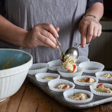 woman in grey scooping strawberry muffin batter from a blue mixing bowl into white paper muffin cases in a grey baking tin