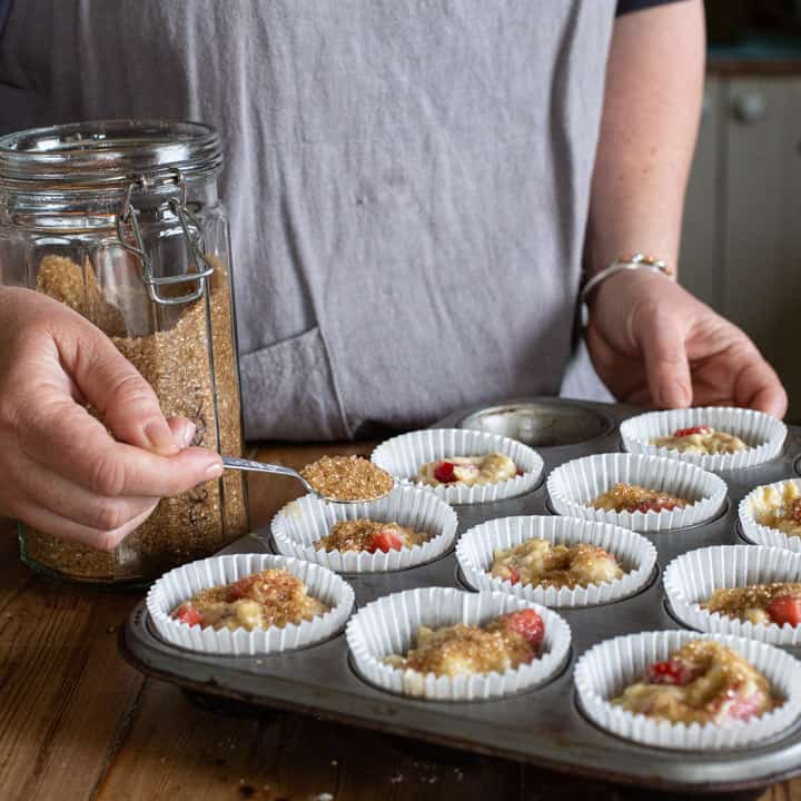 woman in grey sprinkling brown sugar over the top of strawberry muffins about to go into the oven