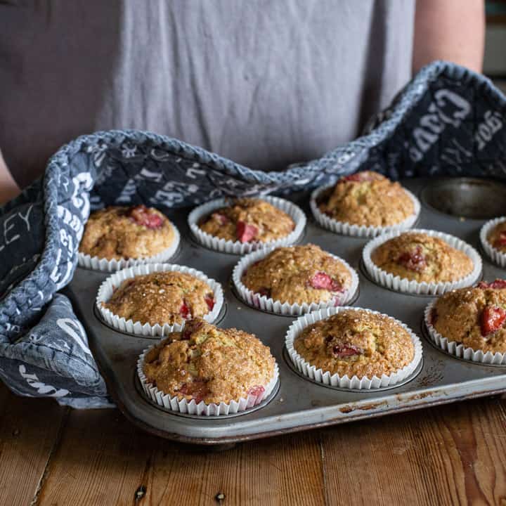 woman in black oven gloves holding a tray of fresh, hot strawberry muffins in a grey muffin tin