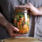 Woman’s hands holding a large glass jar filled with chopped carrots, green coriander and pieces of garlic