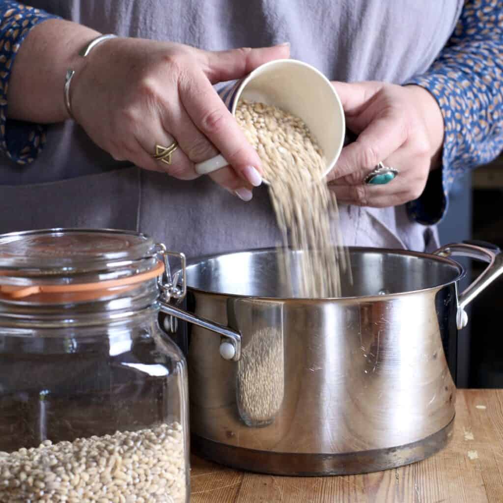 Woman in grey pouring barley from a tea cup into a silver saucepan