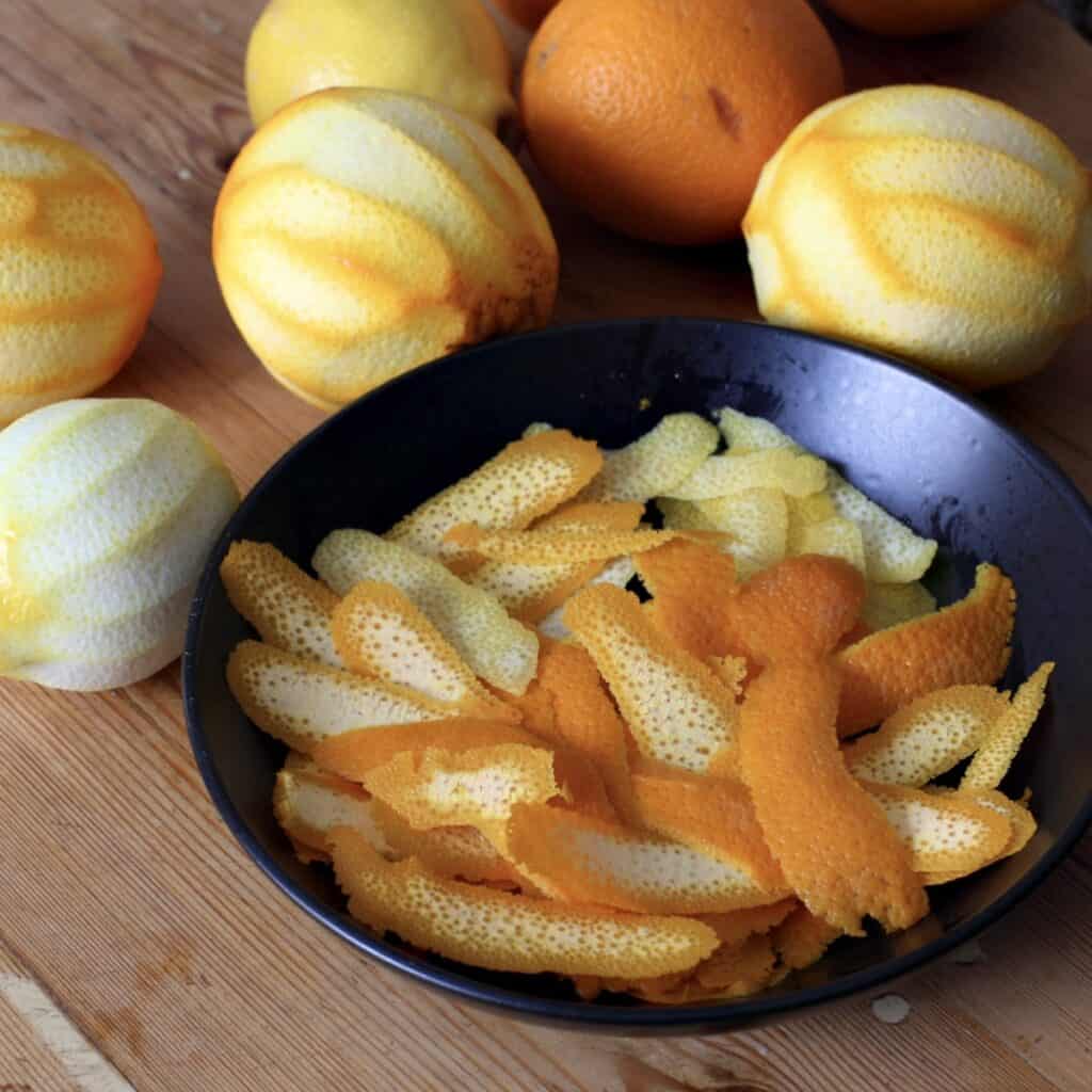 Black bowl holding orange and lemon peelings surrounded by oranges and lemons