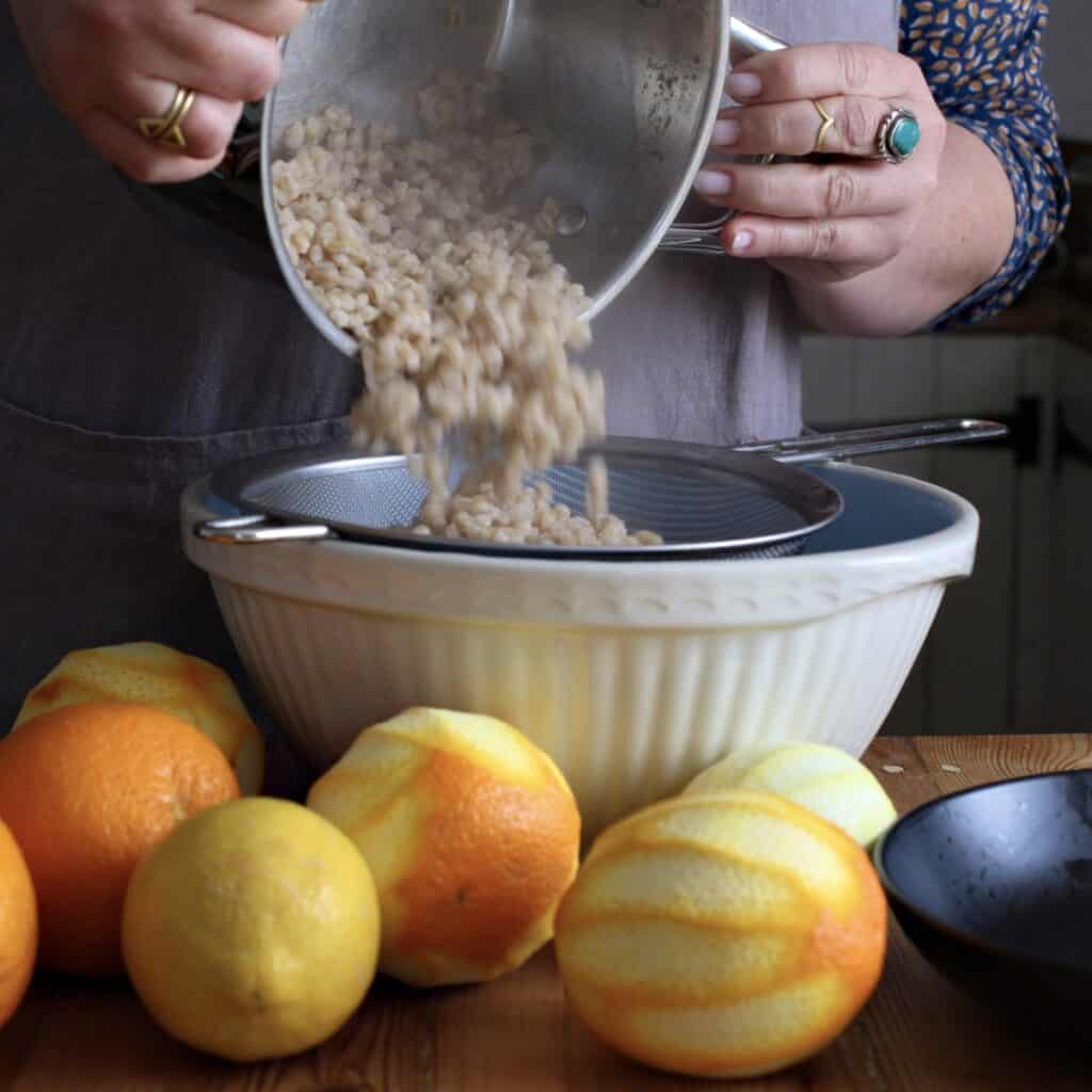 Woman tipping cooked pearl barley from a silver saucepan into a silver sieve balanced over a white mixing bowl