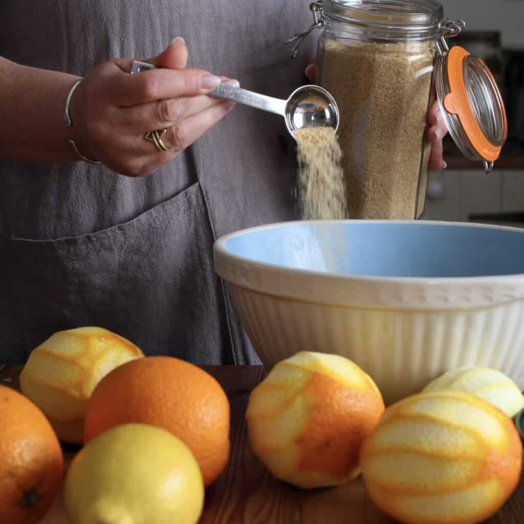 Woman in grey tipping a spoonful of brown sugar into a large mixing bowl with oranges and lemons in the foreground