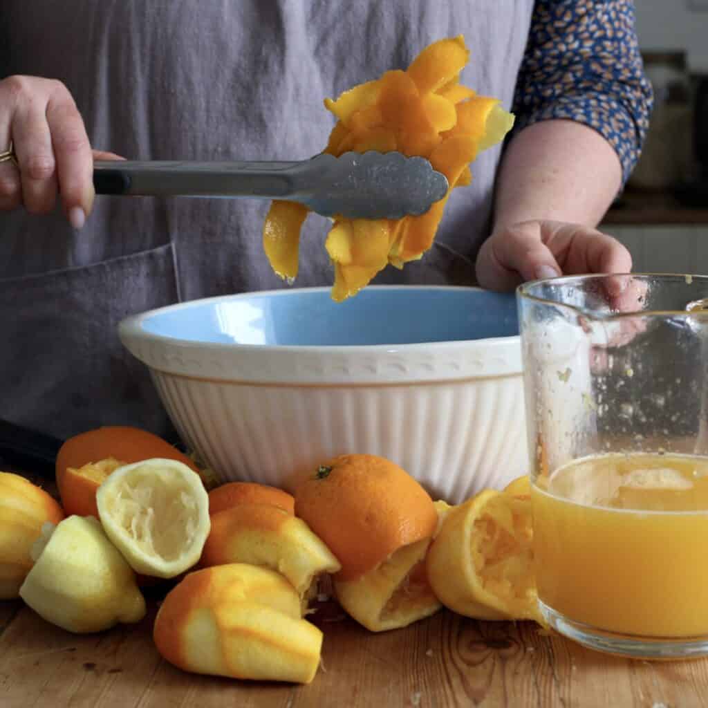 Womans hands removing citrus peels from a blue and white mixing bowl with silver kitchen tongs