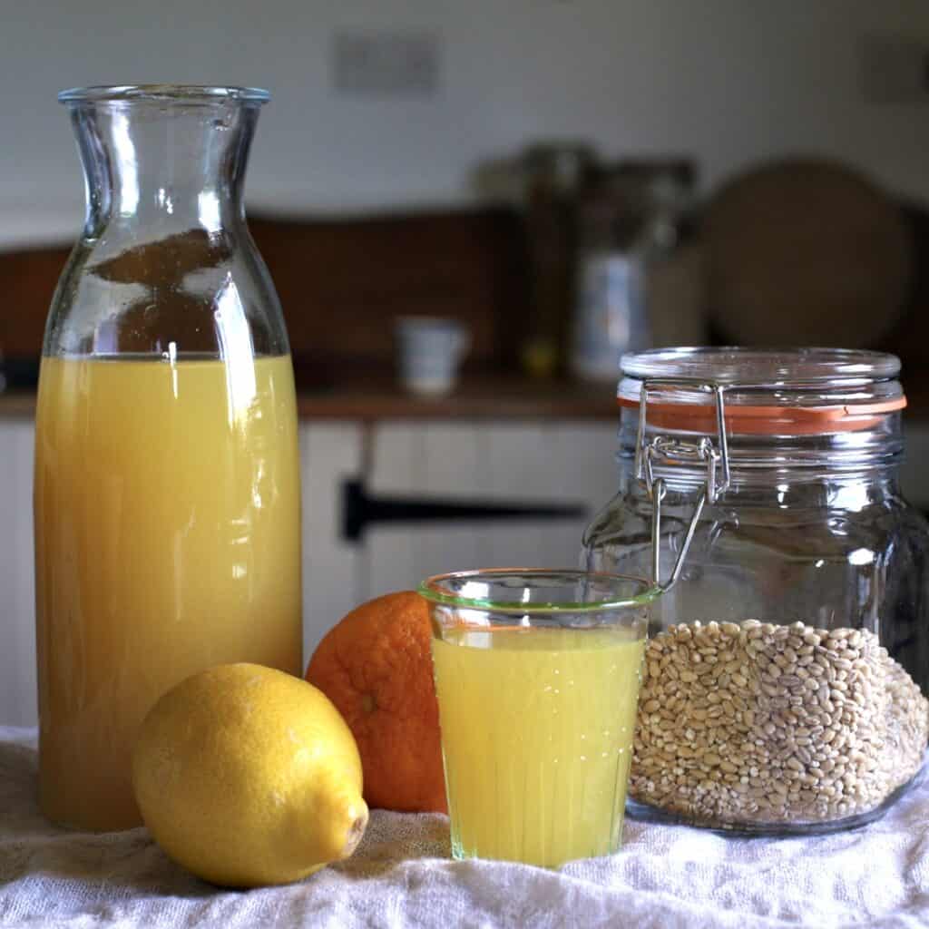 Stilllife on an orange, lemon, a glass and a carafe of orange coloured barley water and a glass jar of pearl barley