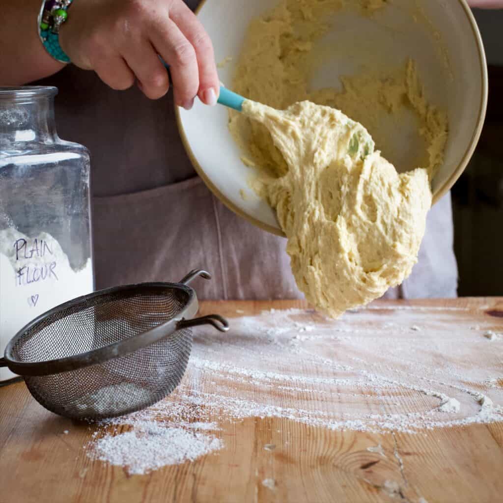 Womans hands scraping cake dough from a white mixing bowl onto a flour dusted kitchen counter