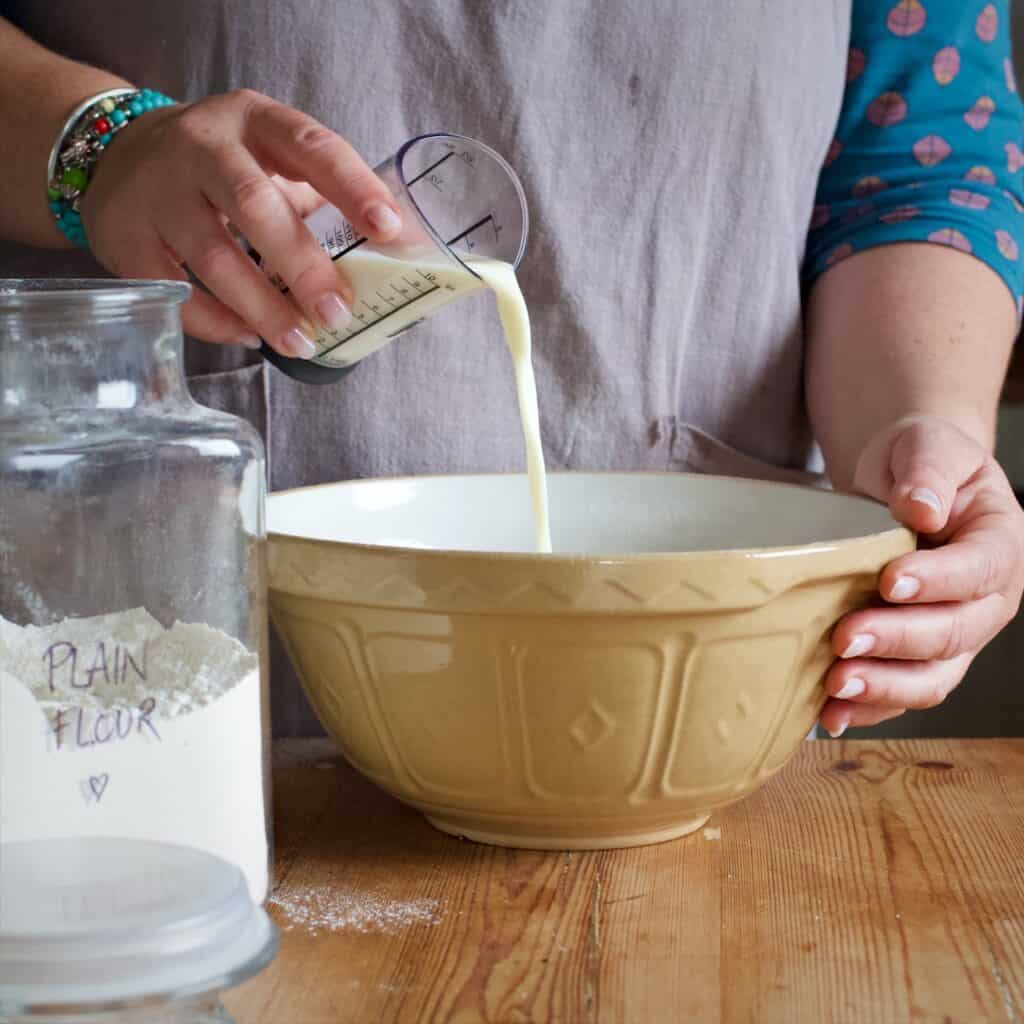 Womans hands pouring milk from a small plastic measuring cup into a large mixing bowl