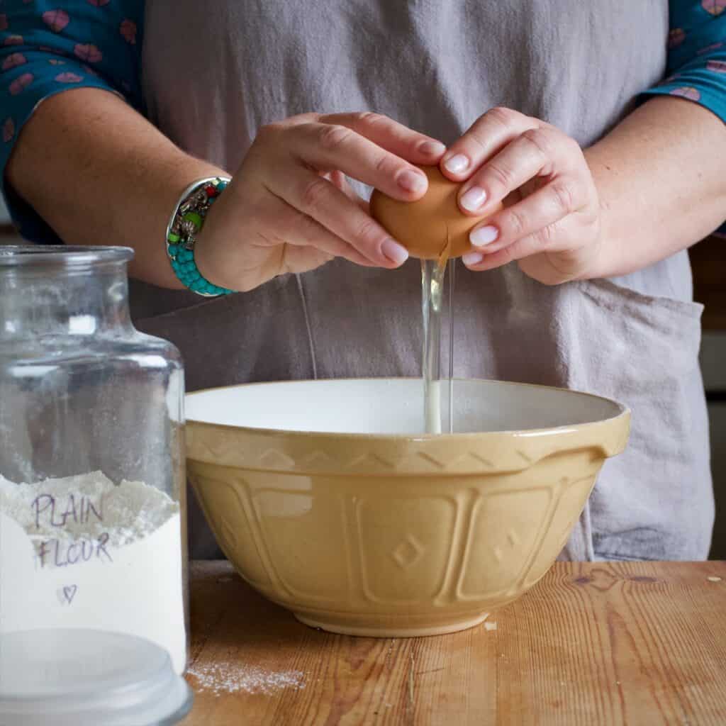 Woman in grey cracking an egg into a brown and white mason cash mixing bowl