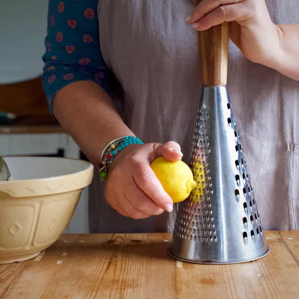 Woman in grey zesting a lemon with a large silver grater on a wooden kitchen counter